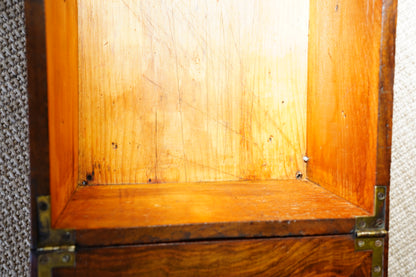 Close-up of the interior of a wooden cabinet or shelf, showing a warm orange-brown wood grain surface with brass corner hardware against a textured neutral background.
