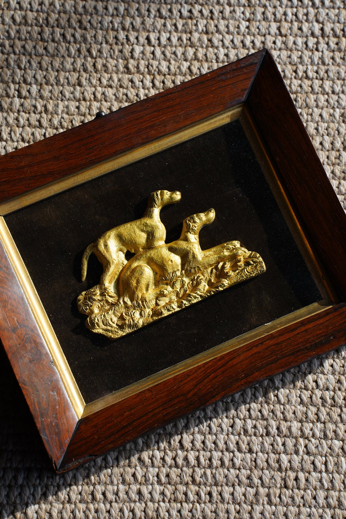 A square framed decorative piece featuring a raised gold figure mounted on a black background, set within a dark wood frame with a gold inner border, photographed on a textured beige surface.