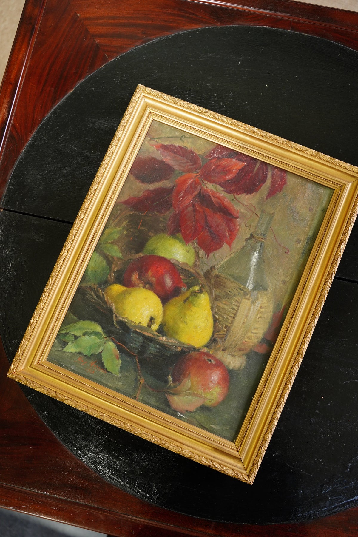 A framed still life painting of red apples, yellow pears, and green leaves, set in an ornate gold frame and displayed on a dark round table.