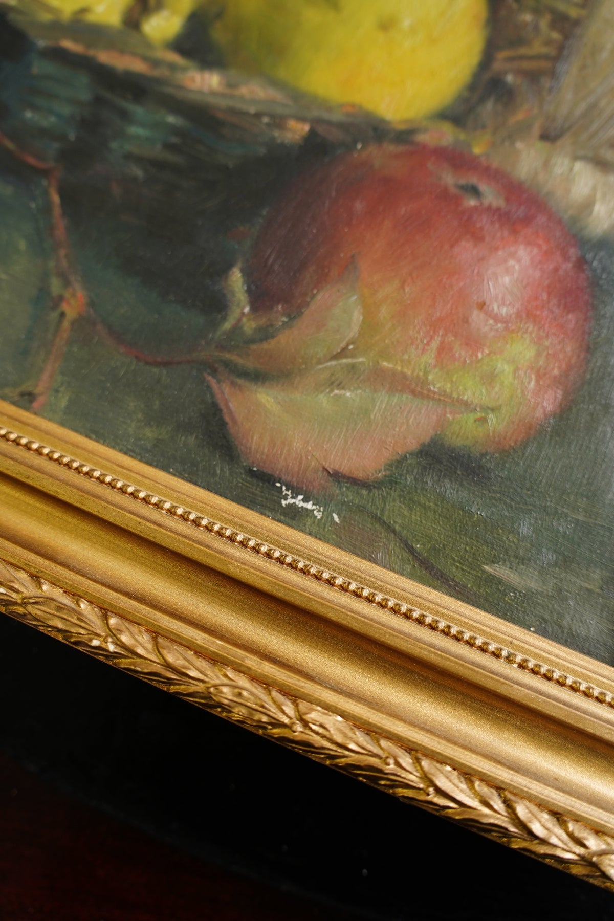 Close-up of a framed still life painting showing a red apple with a yellow fruit at the side, bordered by an ornate gold frame.