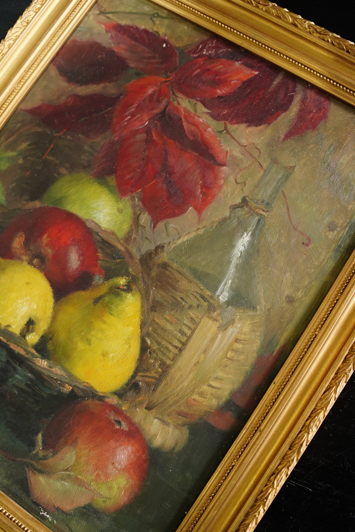 A framed still life oil painting depicting a basket of yellow pears and red apples, accented by red autumn leaves, set in a gold frame.